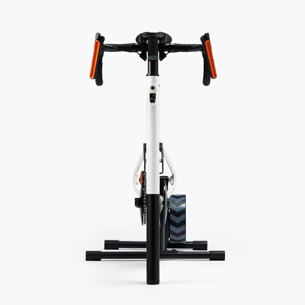 Front view of a stationary exercise bike with a white frame, black handlebars with orange grips, and a black flywheel on a chevron-patterned stand against a plain white background.