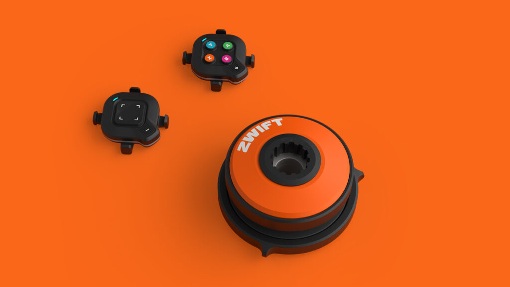 Three black and orange Zwift cycling device components, including a large circular trainer and two small button controllers, are displayed on an orange background.