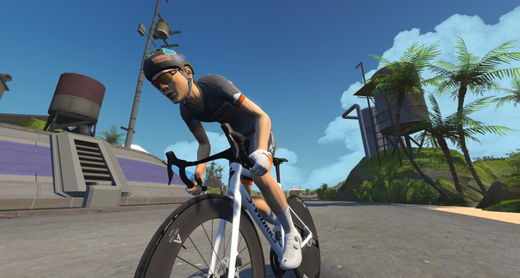 A cyclist wearing a helmet and sunglasses rides a road bike on a sunny day in a virtual environment with palm trees, industrial structures, and a clear blue sky.