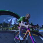 A cyclist in green rides a colorful bike on a road under a large green arch, with mountains, trees, and a house in the background on a clear, sunny day.