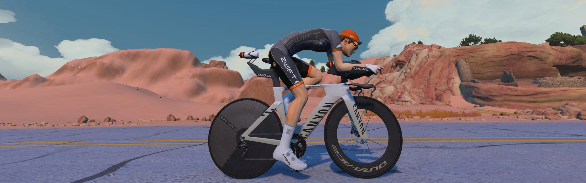 A cyclist in racing gear rides a white Canyon bike on a road through a rocky desert landscape under a partly cloudy sky.