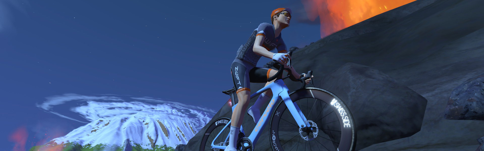 A cyclist wearing a helmet and sports gear rides uphill on a road bike at night, with a snow-capped mountain and a glowing volcano in the background.