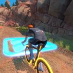A cyclist with an orange helmet rides a yellow bike on a dirt path through a rocky canyon in a virtual cycling game, approaching a glowing blue power-up on the ground.