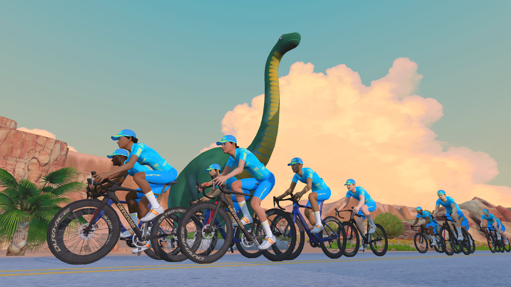 A group of cyclists in blue uniforms ride on a road with a large, green dinosaur statue in the background and a scenic, desert-like landscape.