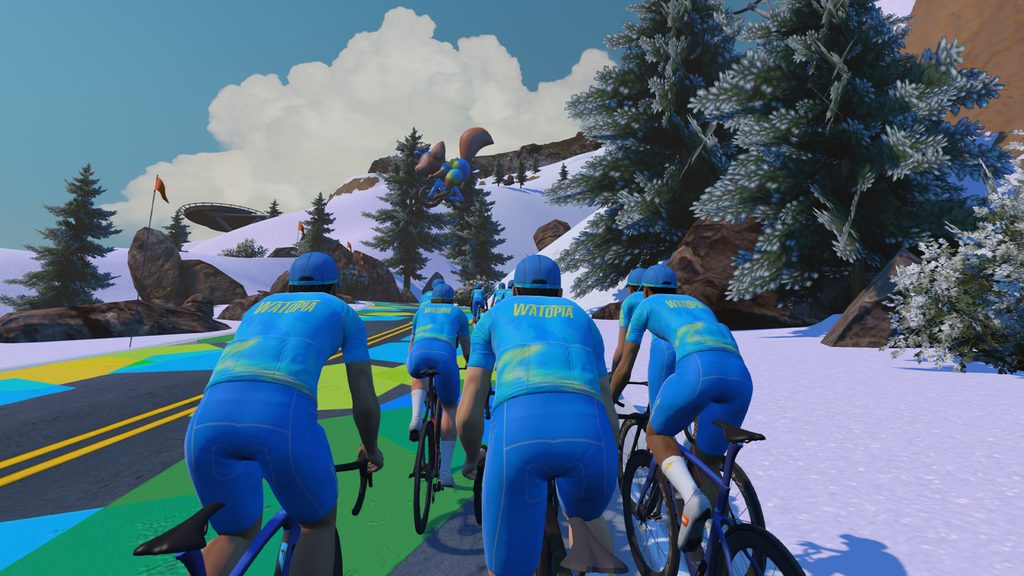 A group of cyclists in matching blue kit ride uphill on a colorful road through a snowy, mountainous landscape with trees and clouds in the background.