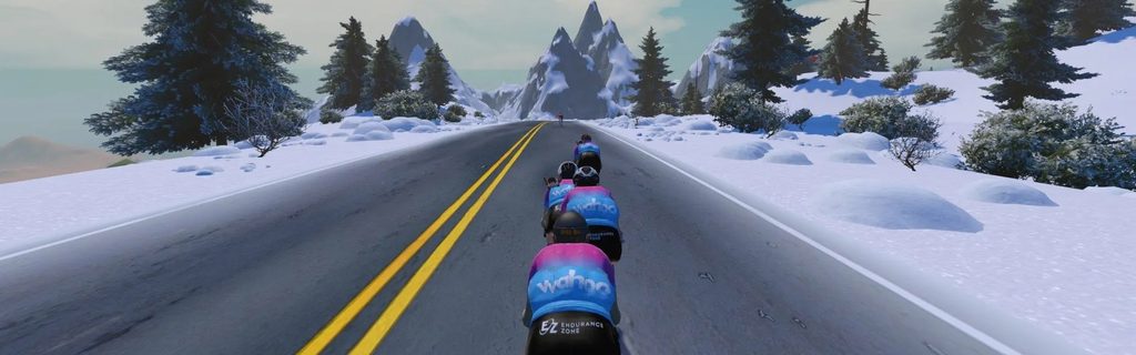 Cyclists in colorful jerseys ride in a line on a snowy mountain road, surrounded by trees and distant peaks under a partly cloudy sky.