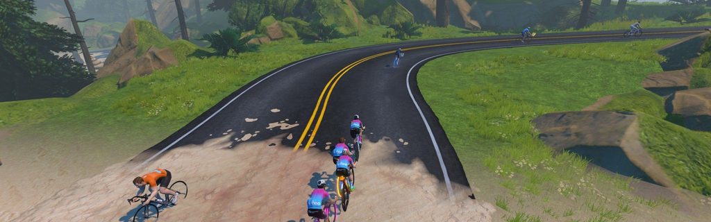 A group of cyclists in colorful jerseys rides on a winding road through lush greenery, with trees and rocks nearby and a few more cyclists visible in the distance.