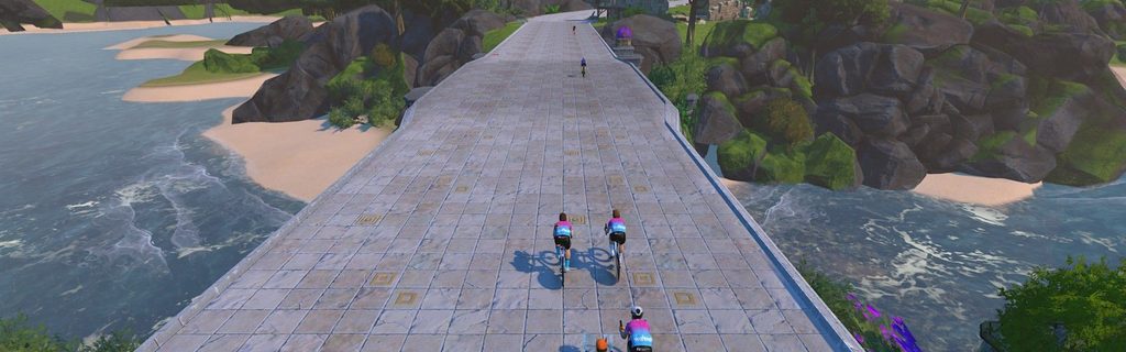 Four cyclists ride along a wide, stone bridge above water and sandy beaches, surrounded by green hills and rocky cliffs under a clear sky.