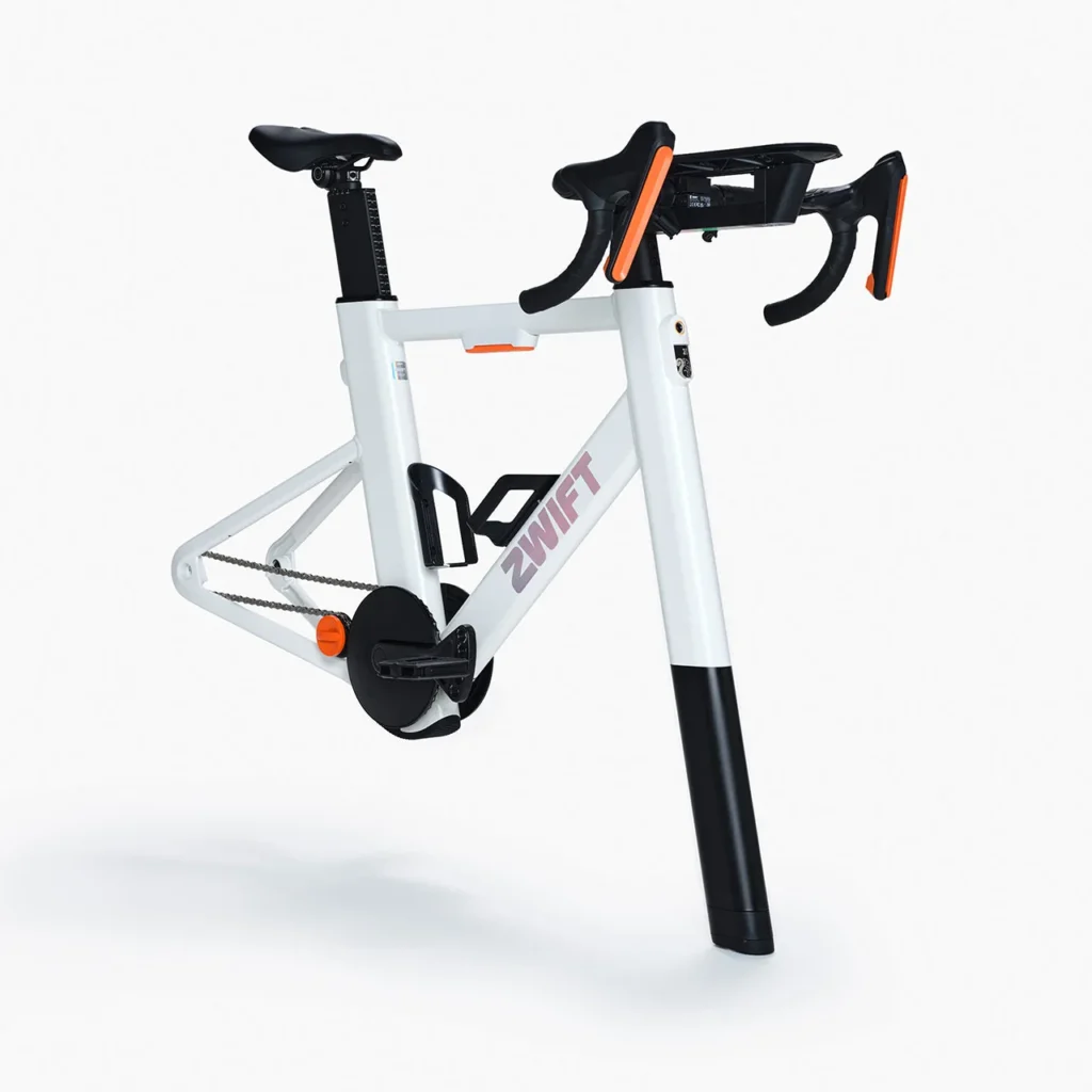 A modern indoor cycling bike with a white frame labeled ZWIFT, orange accents on the knobs and handlebars, and a black flywheel. The bike has no visible wheels and is designed for stationary cycling workouts.