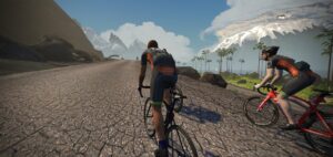 Two cyclists ride road bikes on a cracked paved road through a mountainous landscape.