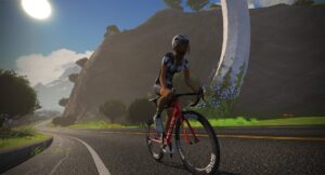 A cyclist wearing a helmet and matching jersey rides a red road bike along a curved road toward a large, curved stone arch.