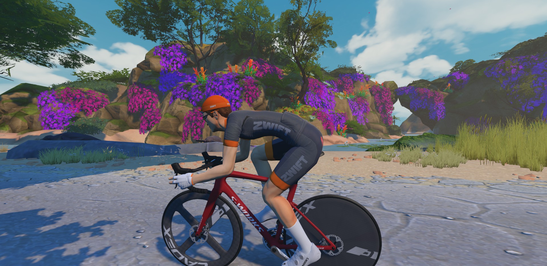 A cyclist wearing a Zwift jersey and orange helmet rides a red racing bike along a path next to a rocky, flower-covered landscape.