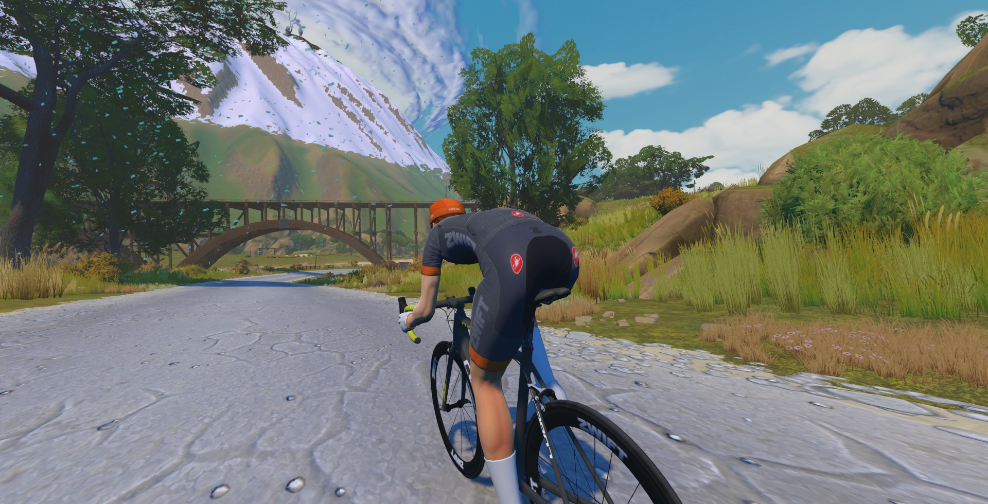 A cyclist in a grey and orange jersey riding a bike on a dirt road toward a wooden bridge with a mountain in the background.