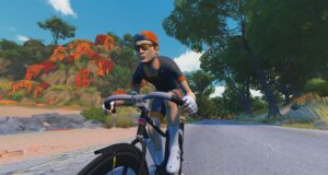 A cyclist in a grey and orange jersey, cap, and sunglasses rides a bike along a coastal road.
