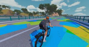 A cyclist in a grey and orange jersey rides a bicycle across a bridge decorated with large blue, green, and yellow geometric shapes.