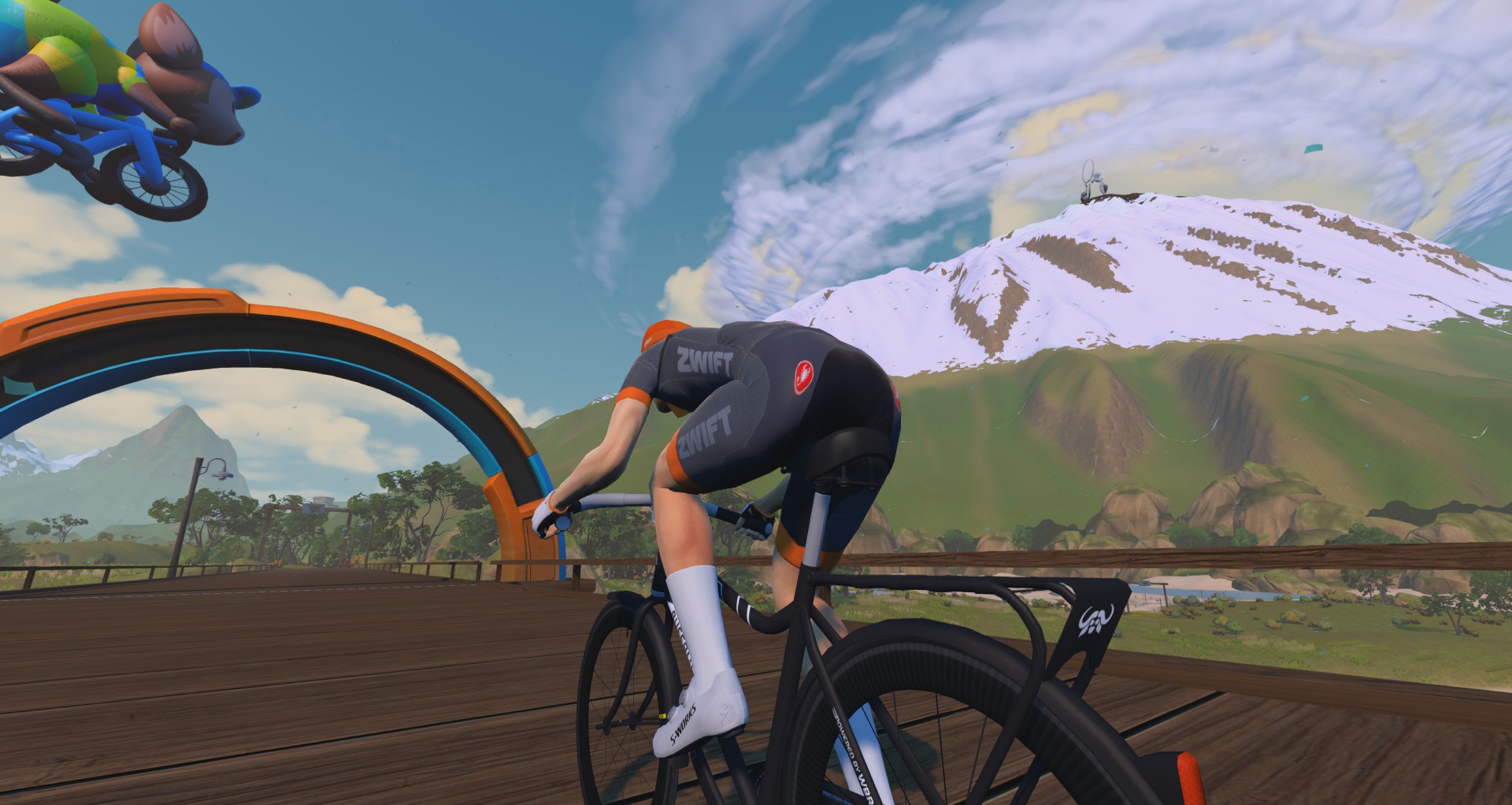 A cyclist riding a bike on a wooden bridge toward a snow-capped mountain, with a second cyclist leaping through the air above.