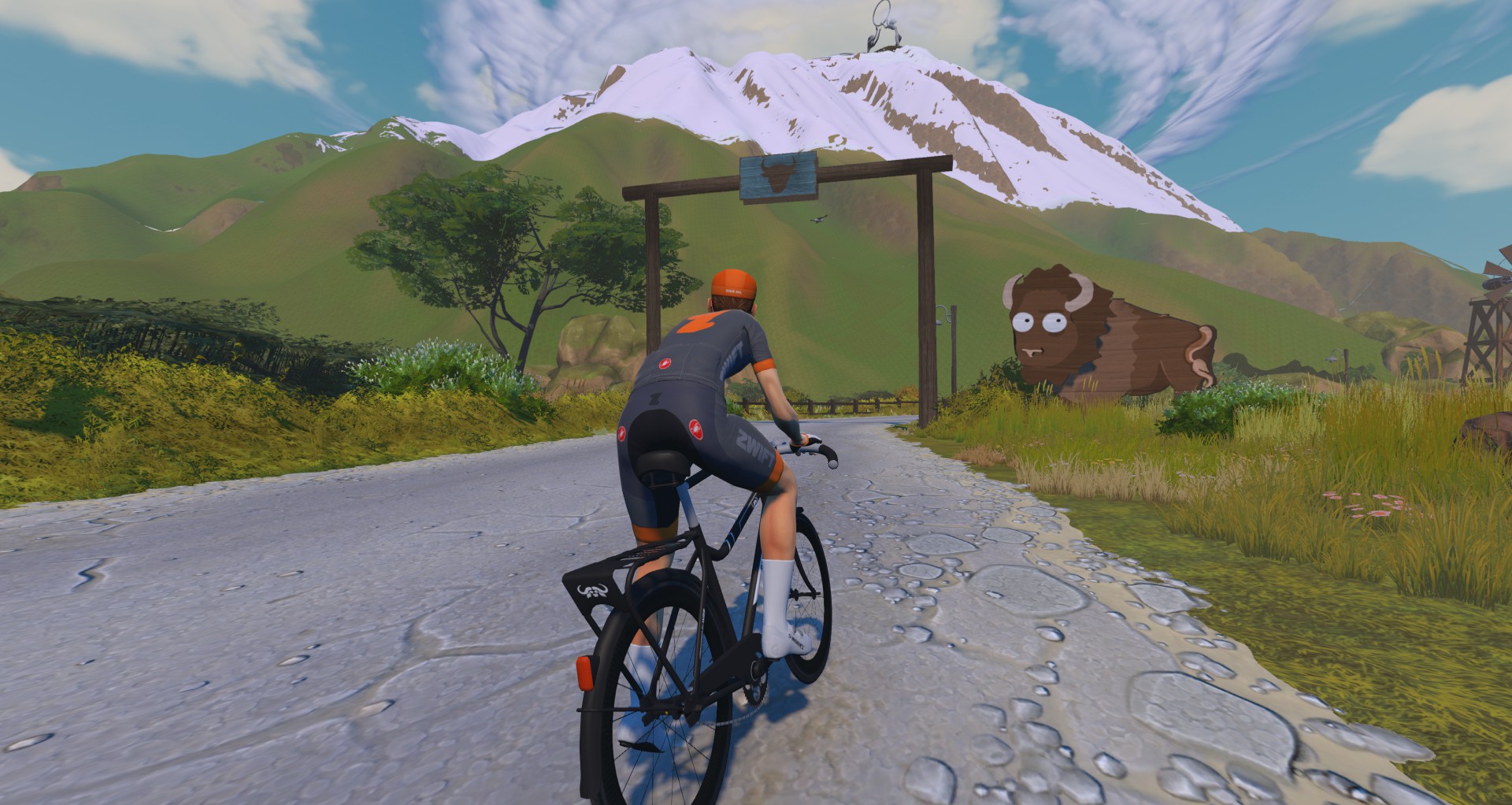 A cyclist in a branded jersey rides a road bike on a gravel path toward a wooden archway with a large bison statue standing to the side, set against a backdrop of mountains.