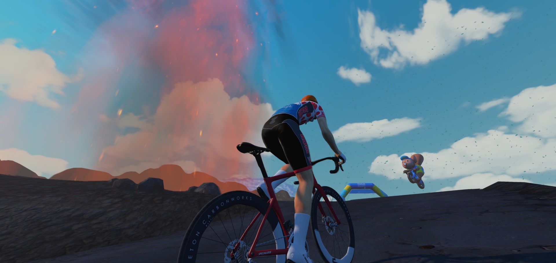 A cyclist rides a red road bike uphill toward a finish arch with an erupting volcano in the background.