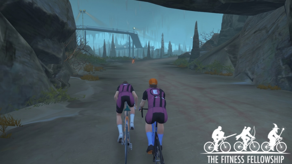 Two cyclists ride through a rocky, misty landscape in a virtual simulation, with the logo for The Fitness Fellowship in the bottom right corner.