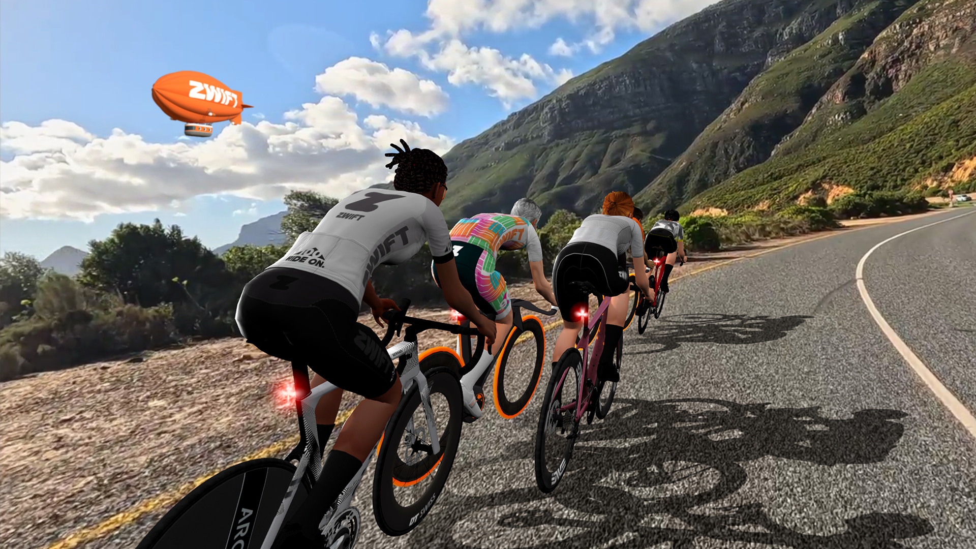A group of four cyclists riding road bikes on a sunny, mountainous coastal highway with a Zwift blimp flying overhead.