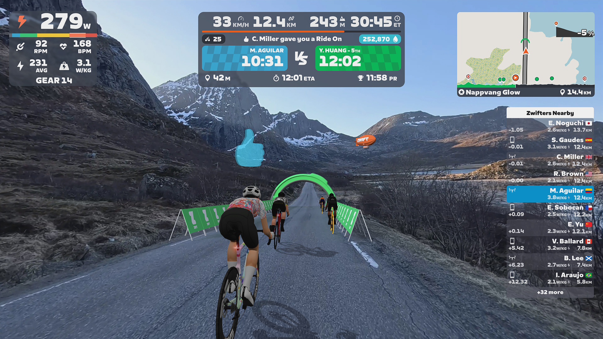 A cyclist riding on a digital road through a mountain landscape, shown from a first-person perspective with a virtual cycling interface displaying speed, power, and other riders nearby.
