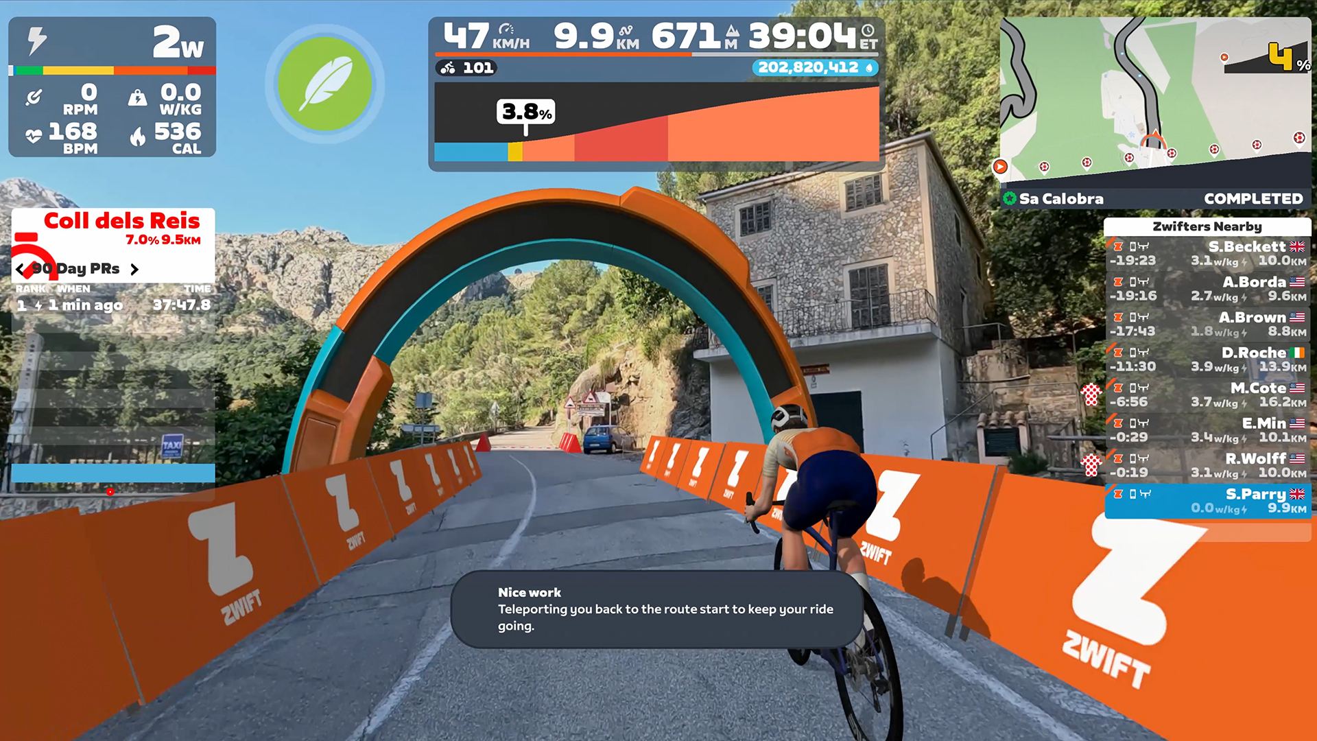 A cyclist rides through an orange Zwift arch on a winding mountain road, shown from a first-person perspective with digital ride metrics and a Zwifters Nearby leaderboard displayed on the screen.