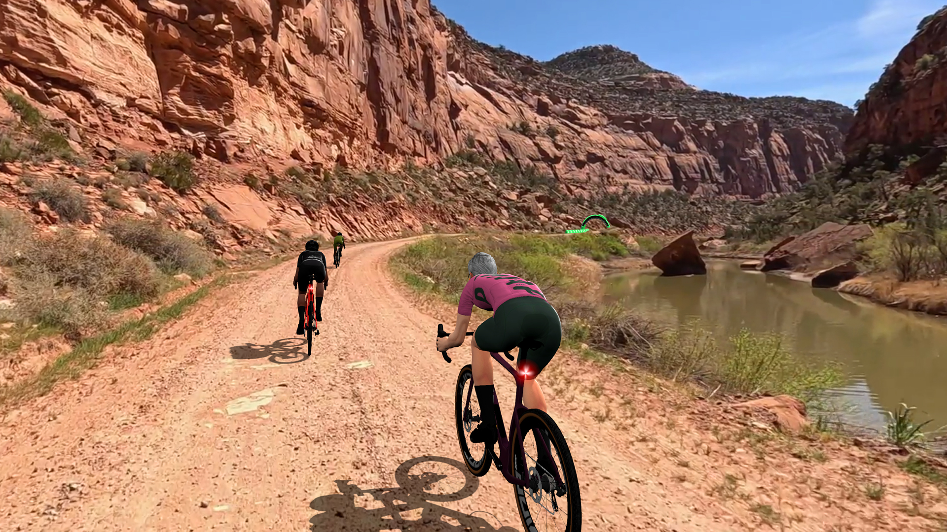 Three cyclists ride road bikes on a dirt trail beside a river at the base of red rock cliffs.