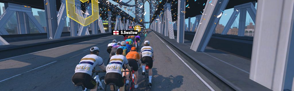 A group of cyclists in a virtual race ride across a large suspension bridge as confetti falls.